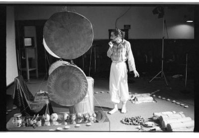 A man wearing a patterned tunic and light pants stands next to two large round hide drums suspended from the ceiling. Various handmade percussion instruments are arranged on the floor and enclosed by a ring of tealights. 