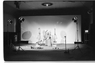 A stage area features a person seated playing various drums: a hand drum is placed between their legs while four round hide drums hang from the ceiling on either side of them. They are encircled by tealights arranged on the floor. There is a large blank backdrop behind, and shadows are cast upon it by large standing stage lights.