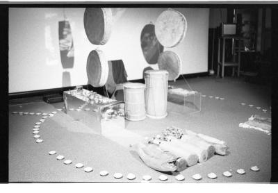 Tea lights are arranged in a circular pattern on the floor. Inside this arrangement are several drums, a chair, and other objects. Four round hide drums hang from the ceiling. 