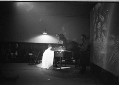 A long-shot image of a man playing grand piano, the front of which is cloaked in a sheet. A guitarist stands to the right of the piano, in front of a screen with a design projected onto it. The pianist is illuminated by a wall sconce.
