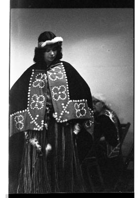 A young woman performs in traditional Kwakiutl regalia. Her gaze shifts down as she dances. An elder is seated behind her as she watches her perform.
