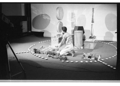 A man sits on his knees at the centre of a stage surrounded by an arrangement of objects and drums within a circular arrangement of tealights on the floor. Four circular drums are suspended from the ceiling.