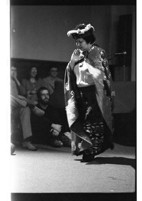 A woman dressed in traditional Kwakiutl regalia performs a dance while an audience in the background observes.