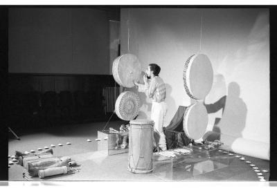 A man stands between two rows of drums hanging parallel to one another, each row having two drums. He places his right hand on the drum while he brings his left hand to his mouth in a whistling gesture. The arrangement is encompassed by a ring of tealights arranged on the floor. There is a large blank backdrop behind him, which is cast with shadows from the drums and person performing. 