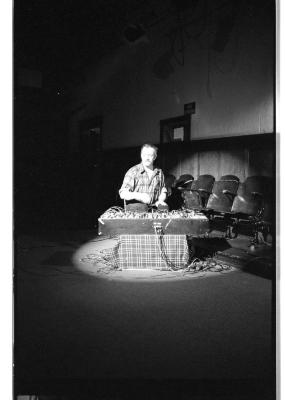 A man in a plaid shirt plays a synthesizer under a spotlight. A row of wooden theatre chairs can be seen behind him. The image is in black and white. 