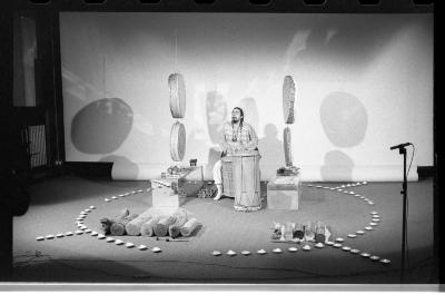 A circular arrangement of tealights on the floor demarcates a performance area. Within it, a man plays a hand drum amongst other objects and instruments. The performer is flanked by two sets of two circular hide drums suspended from the ceiling. 