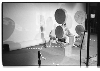 A ¾ long shot of a person seated as they play a drum. They are seated between two rows of drums hanging parallel to one another, each row having two drums. Encompassing them is a ring made up of several small objects, and within it more instruments are arranged. There is a large blank backdrop behind, which is cast with shadows from the drums and person performing. 