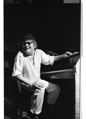 A man wearing a light coloured tunic and pants, round glasses, and a toque is seated at a piano. One of his hands rests on his knee, and the other on top of the piano. Smiling, his gaze shifts to the left of the image. This image is in black and white.