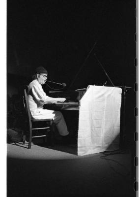 Seen in profile, a man wearing a light coloured tunic and pants, round glasses, and a toque plays grand piano. A piece of cloth is draped over the side of the piano. This image is in black and white.