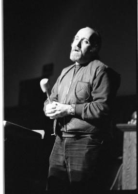 A medium-long shot of a man standing with a microphone in hand. His eyes are partially closed and his mouth is open. There is a stark light cast over him. This image is in black and white.