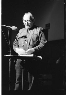 A man wearing round glasses peers down at a book he is holding. Next to him a microphone is propped on a stand and there is a music stand with another book resting upon it. There is a stark light cast over him. This image is in black and white.