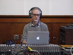 Peter Courtemanche sits by a sound mixing board. He is wearing headphones and looking down at a laptop.