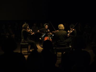 String quartet Quatuor Bozzini sit facing one another in the centre of the Grand Luxe Hall. They are dimly lit in an otherwise dark room as they play viola, cello and violins. Silhouettes of members of the crowd are seen in both foreground and background.