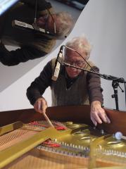 Hank Bull plays the strings of a baby grand piano with a drumstick. A microphone is placed facing the soundboard to capture the sounds. Bull’s reflection is seen in the propped-open lid of the piano.