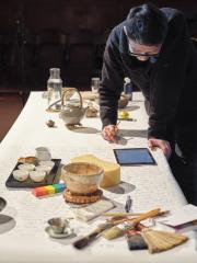 With a pencil in hand, Lam Wong reads off of a digital tablet as he leans on a table covered in a large sheet of paper. There are names scribed halfway down the paper. Also on the table are paint brushes, a book, a sponge, a wooden colour bar, a jug of water, a champagne flute, a singing bowl and assorted tea paraphernalia. 