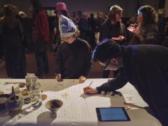 Lam Wong leans over a table covered in a large piece of paper to scribe the names listed on a digital tablet to his left. Wong writes in pencil as a child wearing a newsprint hat stands beside him wielding a marker looks down on his drawing of a creature. Also placed on the table are assorted tea paraphernalia and ceramics, a jug of water, a coaster, a singing bowl, and calligraphy items. Many people converse over tea and cake in the background.