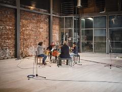 String quartet Quatuor Bozzini sit facing each other in the centre of a live room at The Warehouse Studio. The room, with its wooden floors and walls of brick on one side, and glass on the other, is set up for recording, with several mics placed around the musicians.