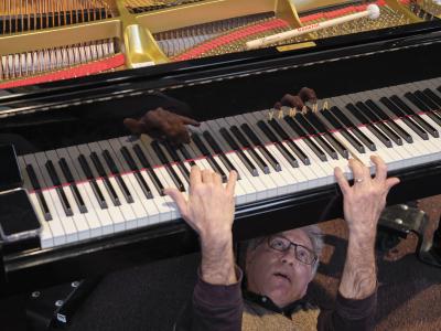 Hank Bull lays beneath a baby grand piano, reaching up from below to play the keys. The piano’s lid is open, revealing a mallet sitting on its tuning pins. 