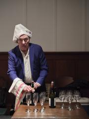 Wearing a blue blazer and a hat made of newsprint, Randy Gledhill stands behind a table set with wine glasses, champagne flutes and wine bottles. With a waiter’s cloth draped on his arm, he prepares to open a bottle of sparkling wine.