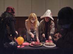 Meghan Latta and Anna Tidlund, who is wearing a newsprint hat, cut into two separate cakes set on a table among bright pink plates, an orange balloon and champagne flutes. Bryan Mulvihill pours hot water from a thermal carafe into a gilded teapot. Beside him stands an audience member eating a piece of cake off a bright pink plate. The pair stand beside a table filled with tea pots, tea cups on a tray, a woven basket and a red tea cosy with dragon embroidery.