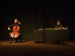 Marina Hasselberg and Sarah Davachi perform in the Grand Luxe Hall. The pair are lit by two spotlights in an otherwise dark room in front of black curtain used as a backdrop. To the left, Hasselberg uses a bow to play the cello. To the right, Davachi adjusts a variety of electroacoustic instruments placed on two tables behind which she sits.