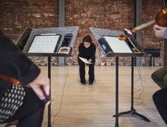 Framed between two music stands, and with a background of a large brick wall, Sarah Davachi sits on the edge of her seat as she looks down at a musical score in a pensive state. The foreground sees music stands equipped with digital tablets showing musical scores and time-keeping devices. Also in the foreground, and slightly out of focus are the right side of Isabelle Bozzini playing the cello and the left side of Alyssa Cheung playing the violin.
