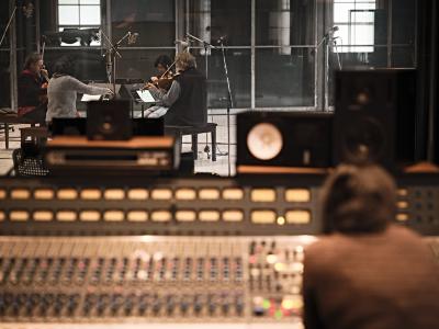 A look through the window of a control room out into a live room at The Warehouse Studio. String quartet Quatuor Bozzini sit facing each other while playing their instruments in the live room, with several microphones placed around the room. In the foreground, Sarah Davachi sits at a mixing desk in the control room.