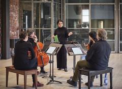 Sarah Davachi addresses string quartet Quatuor Bozzini as they sit in front of their music stands while holding their instruments. They are in a live room at The Warehouse Studio, which has tall ceilings, red bring walls, and another wall made of several panes of glass.