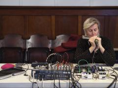 Anna Friz plays a harmonica into a microphone connected to an assortment of electronics and a mixing board sitting on a table.