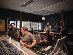 Sarah Davachi and sound engineer Zack Blackstone in front of a large mixing desk in a control room at The Warehouse Studio. String quartet Quatuor Bozzini and Western Front staff members listen in from the back of the room.