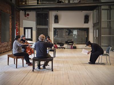 String quartet Quatuor Bozzini and composer Sarah Davachi rehearse in a live room at The Warehouse Studio. The quartet play their instruments while sitting on piano benches in a semicircle, as Davachi faces them, sitting on the edge of her seat pensively looking down towards the musical score in her hands. At the far end of the room is a semi-reflective window looking into the control room.