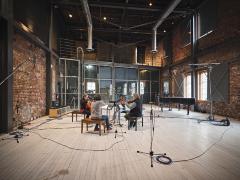 A wide angle shot of a live room at the Warehouse Studio set up for the recording of string quartet Quatuor Bozzini. The room has tall ceilings, two red brick walls, and at the far end, a balcony atop a wall of windows, which overlooks the studio. The quartet sit facing each other in the centre of the room playing their instruments.