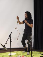 Holding bells attached to strings in both hands, Igor Santizo stands on a green blanket in front of a white screen. On the blanket are several percussive instruments and two standing microphones.