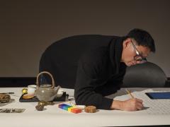 Lam Wong leans over a large piece of paper on a tabletop to scribe names in pencil as part of a performance. Also on the table is a digital tablet, a wooden round, a wooden colour bar, a singing bowl, a photograph, and tea paraphernalia. 