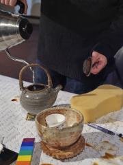 A close up shot of Lam Wong pouring water into a ceramic teapot. The teapot is sitting on a tabletop covered with a tea-stained paper on which several names have been scribed in pencil. Also on the table is a stack of tea cups sitting on a coaster, a sponge, a pen and a wooden colour bar. 