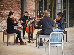 In a circle formed of piano benches and music stands, string quartet Quatuor Bozzin sit facing each other while playing their instruments. They are in the centre of a large room with brick walls. A vacant chair sits in the foreground.