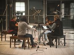 String quartet Quatuor Bozzini sit facing each other in the centre of a live room at The Warehouse Studio. There are several microphones placed around the group as they play their instruments while reading a musical score off of digital tablets placed on music stands. In the background is a wall made of several sections of square glass planes.
