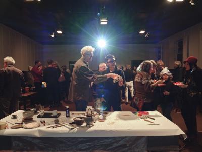 Many people converse over tea and cake in a large hall. The light of a projector shines from the far end of the hall towards the camera. In the centre of the image, two people examine a photograph in front of a table covered in a large sheet of paper on which many names have been scribed in pencil. Als on the table is assorted tea paraphernalia, a champagne flute, a jug of water, several paint brushes, ceramic bowls, a wooden colour bar, a sponge, a bottle of ink. a coaster, a pencil and assorted calligraphy items.