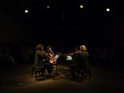 String quartet Quatuor Bozzini sit facing one another in the centre of the Grand Luxe Hall. They are lit by four spotlights overhead in an otherwise dark room as they play viola, cello and violins.