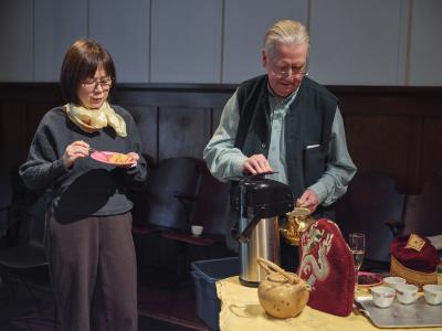 Bryan Mulvihill pours hot water from a thermal carafe into a gilded teapot. Beside him stands an audience member eating a piece of cake off a bright pink plate. The pair stand beside a table filled with tea pots, tea cups on a tray, a woven basket and a red tea cosy with dragon embroidery.