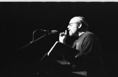 A man seen in profile talks into a microphone. His one hand points upward with his index finger and the other hand holds an open book. Stark lighting is cast upon his face and chest while the microphone, the book, and his body fade into the dark background. This image is in black and white.