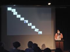 Brand Bird stands behind a lectern affixed with a microphone in the Grand Luxe Hall. Their gaze is cast down as they read from loose sheets of papers held in their left hand. Bird is positioned in front of a projected image by Christian Vistan. The abstract composition shows a line drawing evoking links of a chain cascading across ten white squares that descend diagonally across a black surface. 
