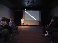 Holding a cordless microphone in her right hand, Kiel Torres reads from a piece of paper in her left hand while addressing the audience in the Grand Luxe Hall. She is positioned in front of a projected image by Christian Vistan. The abstract composition shows a line drawing evoking links of a chain cascading across ten white squares that descend diagonally across a black surface. 