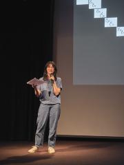 Jacquelyn Zong-Li Ross stands holding a cordless microphone with her left hand and a piece of printer paper in her right hand while addressing the audience in the Grand Luxe Hall. She is positioned in front of a projected image by Christian Vistan. The abstract composition shows a line drawing evoking links of a chain cascading across ten white squares that descend diagonally across a black surface. 