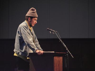 Billy-Ray Belcourt stands behind a lectern in the Grand Luxe Hall. He grips the edges of the lectern while leaning into the affixed microphone.