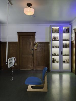 A small monitor suspended from the ceiling faces a chair upholstered in blue fabric. Above the chair, on the ceiling, is a light red light. In the background, pressed against a wood panelled wall, sits a shelf containing books and various ceramic wares.