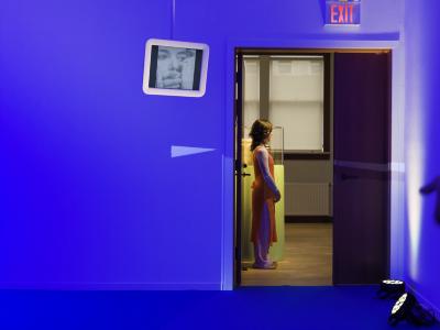 An exhibition guide wearing an orange apron stands outside the open doors of a gallery. Inside, the gallery walls are cast in blue light, and its floors covered in blue carpet. A small, white framed monitor is suspended from the ceiling, showing a black and white image of a person having their mouth covered by several hands.