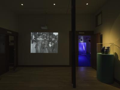 A view from a lobby in which a black and white video of a nude figure trying on leopard print clothing is projected on the wall. To the right, a vitrine containing four vases is lit under a spotlight. An open door offers a peak into a blue-lit gallery space.