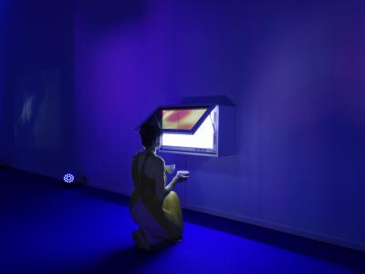 An exhibition tour guide, dressed in a yellow apron, kneels in front of a digital-screened display case, with two tea bowls in hand. The room, covered in blue carpet, is cast in blue light. A video is projected onto the far wall.