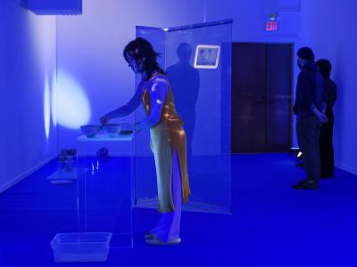 An exhibition guide wearing an orange apron, blue pants, and sandals, prepares for a tea service. She stands over ceramic wears placed on a plexiglass plinth. The room is cast in blue light and various props and lights are placed on the blue carpet. In the background, patrons watch a video on a small monitor.